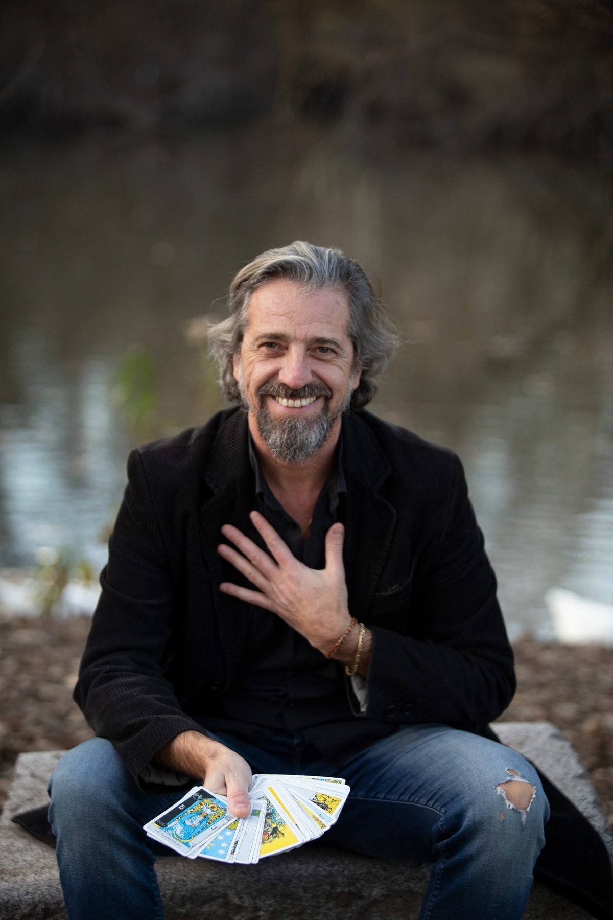 Dario Nencini seduto in riva al lago, sorride con un ventaglio di tarocchi in una mano e l'altra sul cuore